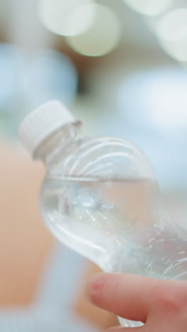 una mujer sostiene agua en botella fría en un centro comercial detrás de otra mujer, sacudiendo gradualmente la botella, el fondo presenta iluminación borrosa, y la botella es clara y fría con condensación visible