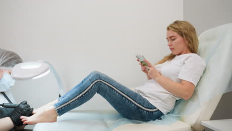 client seated on recliner concentrating on smartphone while receiving pedicure from nail technician, showing casual posture with legs stretched and serene expression in bright salon setting