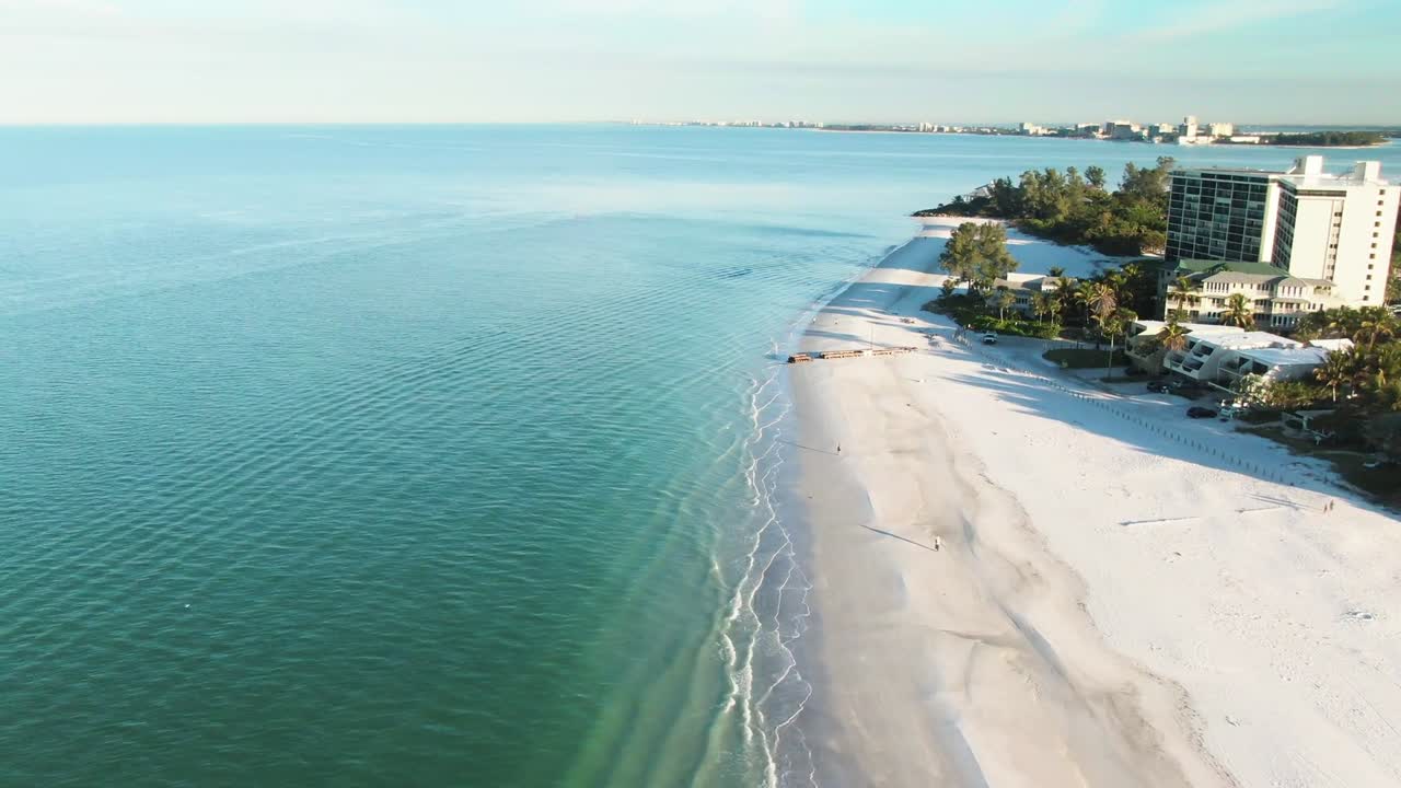 An aerial view of a pristine beach with turquoise waters and gentle waves lapping the shore. A small cluster of buildings sits near the shoreline, surrounded by trees, creating a peaceful coastal view