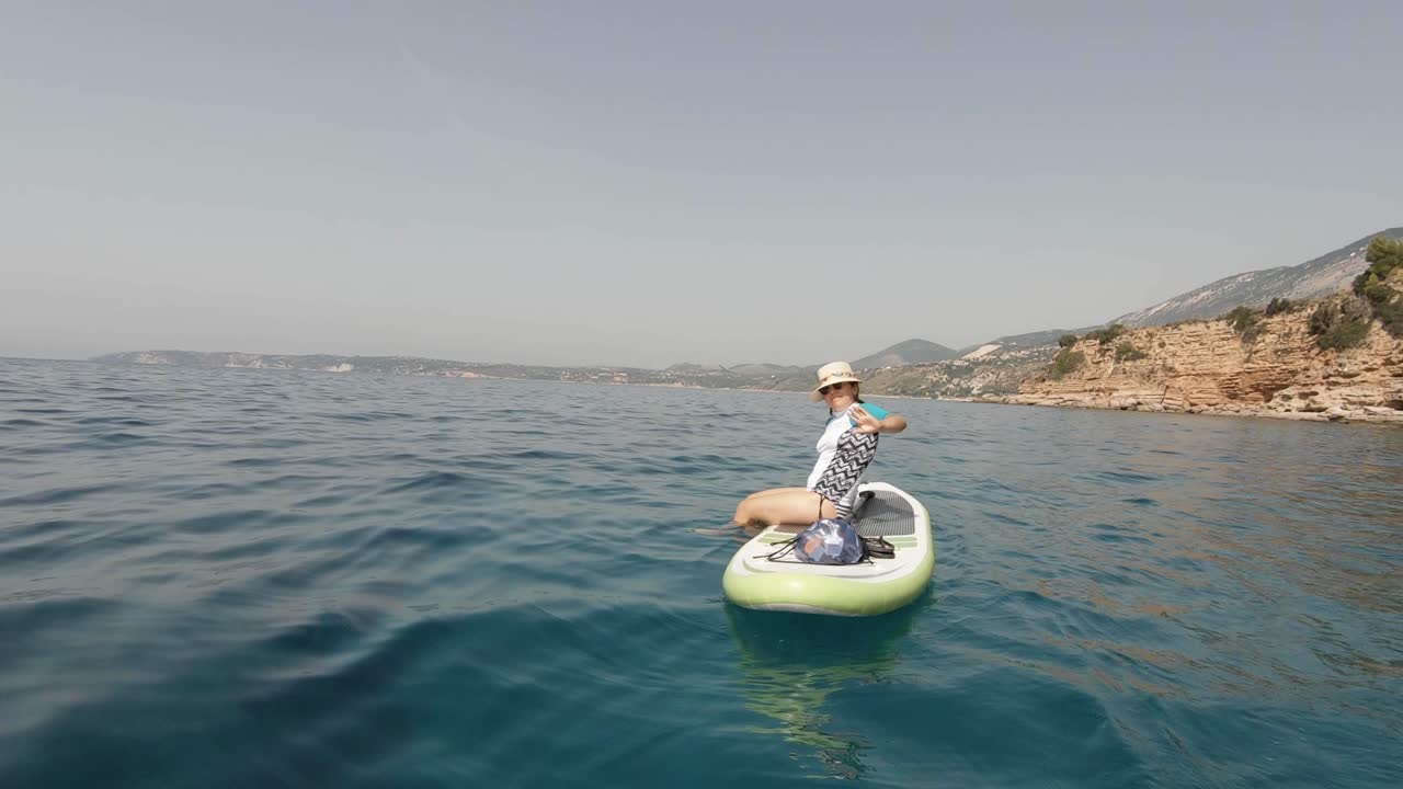 una chica deportiva saludando a la cámara, usando un sombrero y gafas de sol mientras descansa en una tabla de remo de pie