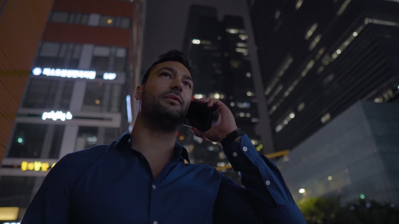 Young businessman is having a phone talk while standing by high-rise skyscrapers in city downtown at night