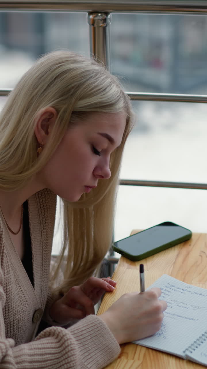 Young lady writes thoughtfully, pauses to place hand on jaw, continues writing with phone and coffee on the table, background features blurred winter scene through window