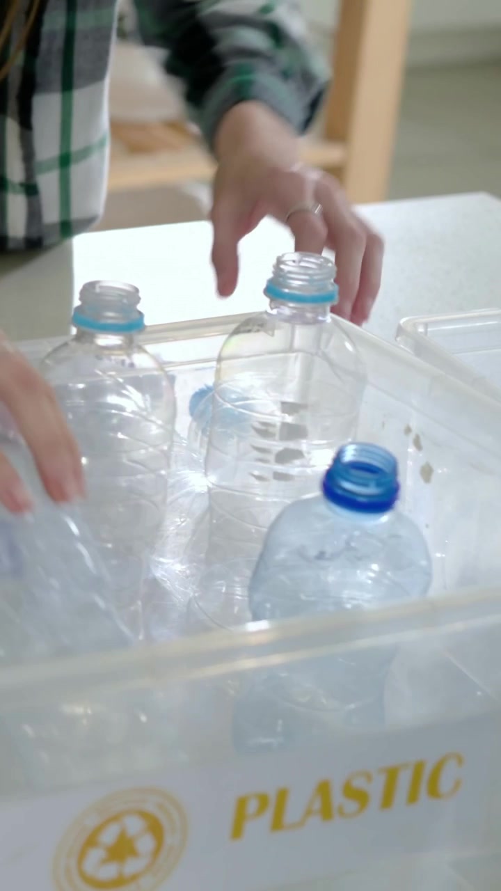 Video verticale: Donna in top a quadri che si protende, smista bottiglie in cucina in un contenitore di PLASTICA, spazio per il testo.
