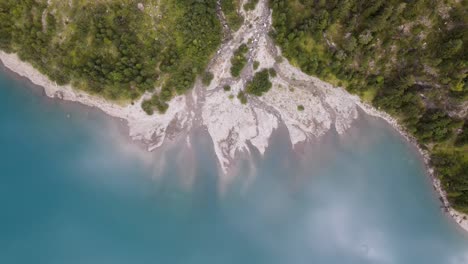 Flying-up-with-slow-spin-over-stream-flowing-into-blue-lake-with-rock-shoreline-and-forest