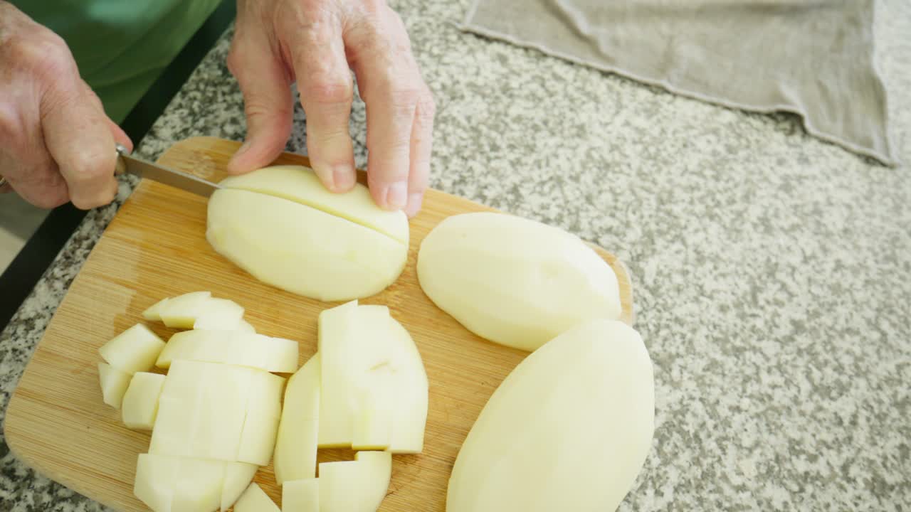 Mani anziane preparano patate a pezzi con un coltello su un tagliere in preparazione per un pasto. Primo piano di mani anziane che preparano un pasto a casa.