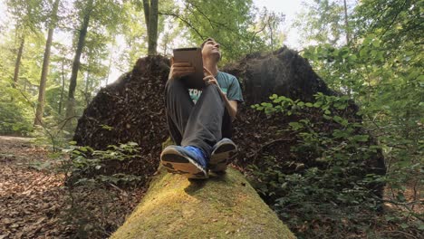 a man is working at his tablet while he's sitting at a mossy trunk in a green forest - business working at the fresh air, modern technology