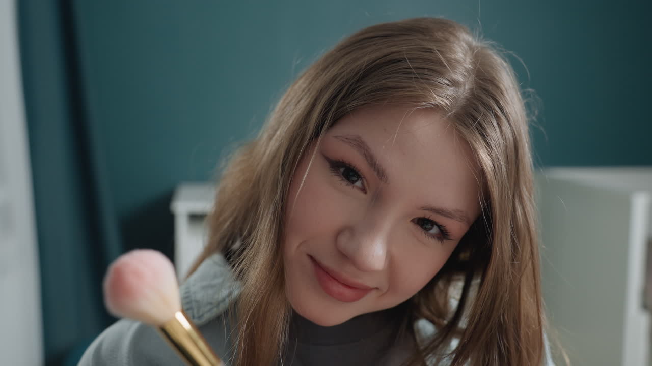 Caucasian girl gentle brush closeup, soft ASMR vibes, delicate strokes over cheek, serene gaze, bedroom backdrop with teal wall, warm lamp glow, subtle glitter falling, calming beauty ritual