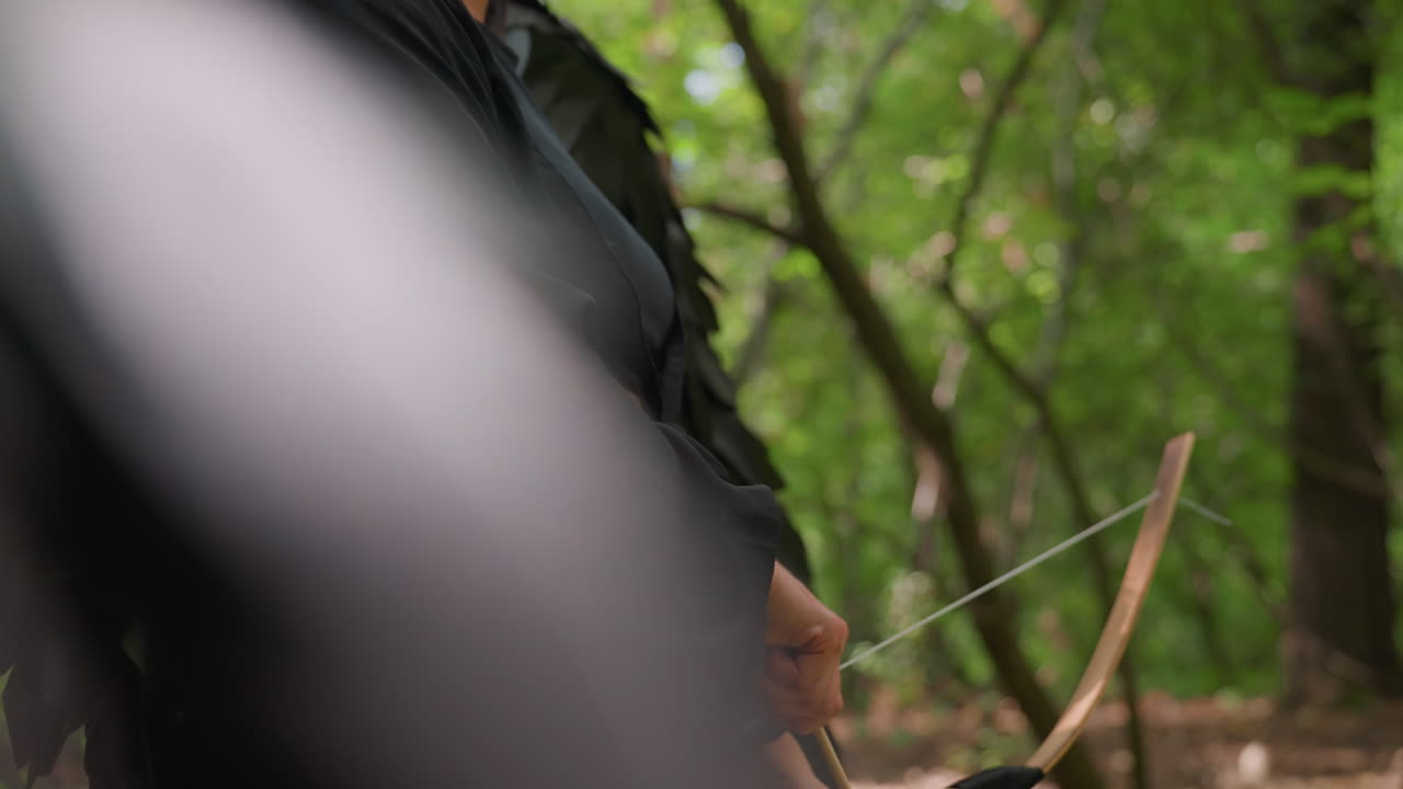 Side view of female archer with black wings standing in sunlight within green forest, bow lowered as she prepares to shoot, expression calm and focused, atmosphere blending power