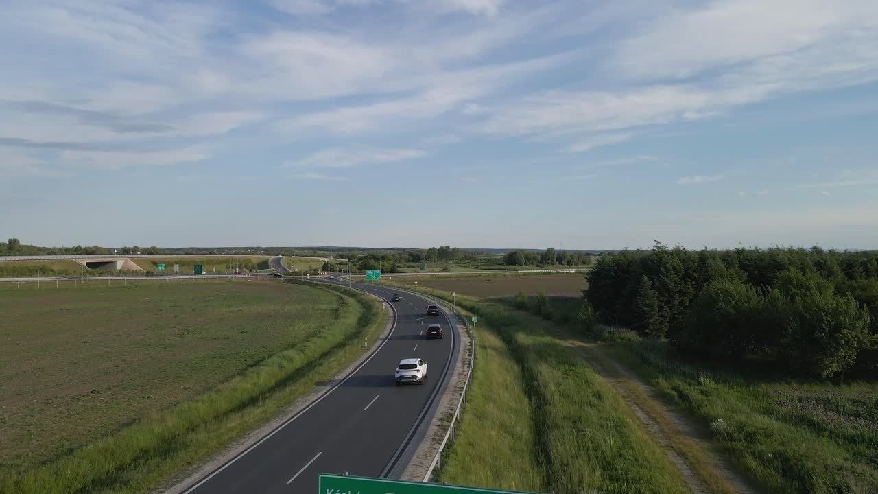 A rural Hungarian highway scene featuring road signs indicating directions to Kópháza, Bécs-Wien, and Győr. Few cars drive along the asphalt road surrounded by fields, trees, and a bright blue sky.