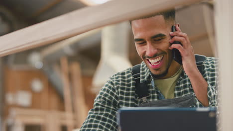 aprendiz masculino con tableta digital trabajando como carpintero en un taller de muebles hablando por teléfono móvil
