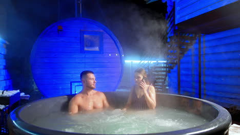 happy caucasian couple sits relaxing in the hot tub. woman in stripy swimsuit speaks on the phone.