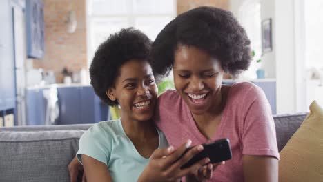 Happy-african-american-mother-and-daughter-sitting-on-sofa-using-smartphone