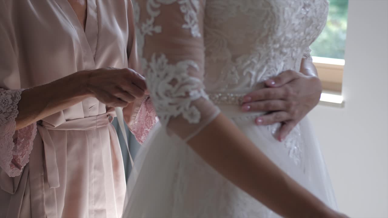 Mother in pastel morning robe helps her daughter put on wedding dress, slowmo