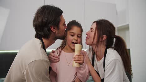 Close-up-of-a-happy-brunette-girl-in-pink-clothes-jumping-and-having-fun-with-her-parents-who-kiss-her-on-the-cheek-while-preparing-for-breakfast-in-the-morning-in-a-modern-apartment-in-the-kitchen