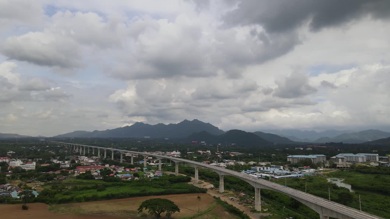 Aerial footage towards as town, Muak Klek, in Saraburi, Thailand, revealing an elevated railway on the right and mountains in the horizon with rain clouds