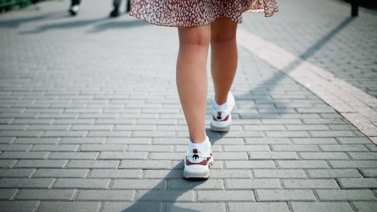 Vista delle gambe di una donna che cammina con un vestito a fantasia vintage e sneakers di tela bianche su un sentiero pavimentato, con le ombre di altre persone che si avvicinano dalla direzione opposta, proiettando ombre sul sentiero illuminato dal sole