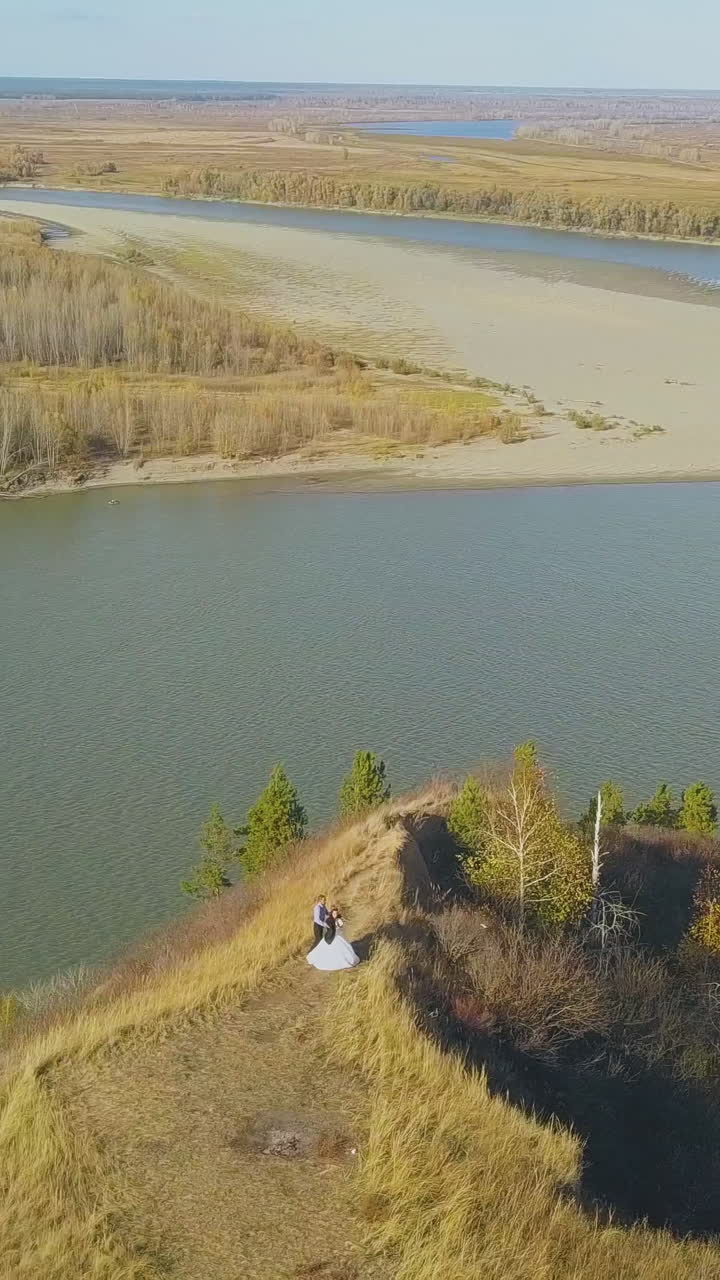 just married couple looks at calm river from steep bank with dry grass against pictorial landscape on autumn day aerial