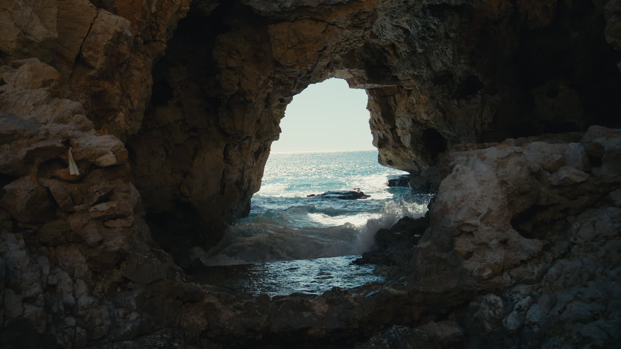 Muri di pietra erosi incorniciano una luminosa apertura verso il mare mentre le onde si spingono nella grotta, creando una scena costiera selvaggia con contrasto, movimento e un tranquillo fascino.