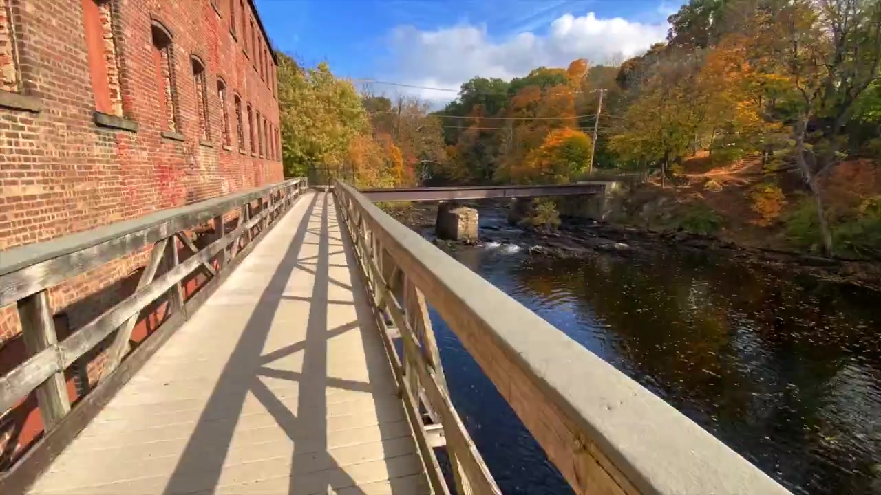 Timelapse af en gåtur langs en gangbro ved en bæk om efteråret i et forladt industriområde, med en bro, rindende vand og farverigt løv