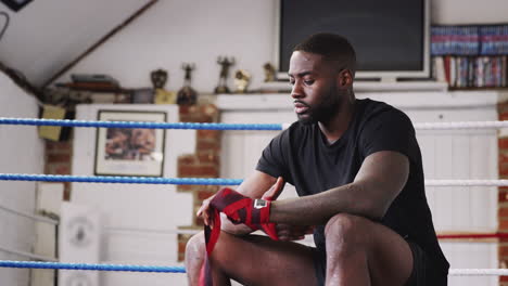 Männlicher-Boxer-Trainiert-Im-Fitnessstudio,-Sitzt-Im-Boxring-Und-Legt-Wraps-Auf-Die-Hände