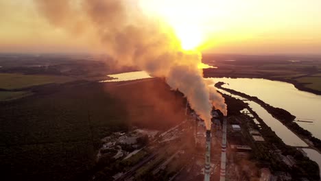 pipes with smoke on the natural green fields background. clouds of smoke coming from the chimneys of the plant with yellow lighting of setting sun. aerial view.