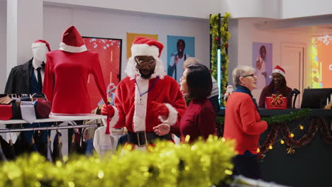 employee wearing santa claus costume around shopping mall clothing shop to spread holiday joy. worker in festive themed suit showing happy client red garment piece during winter holiday season