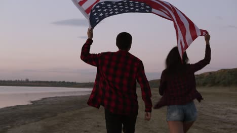 Young-couple-both-in-red-plaid-shirts-are-running-on-the-seashore-with-american-flag-raised-above-their-heads.-Backside-view