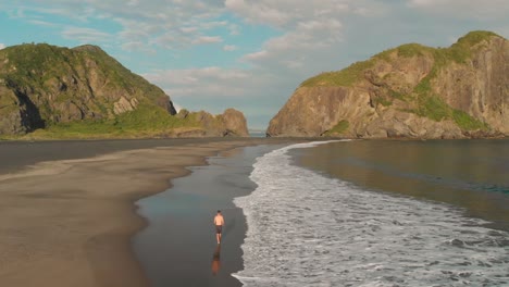 slowmo - aerial drone shot of young man walking on black sand vulanic whatipu beach by ocean towards incredible rock formations