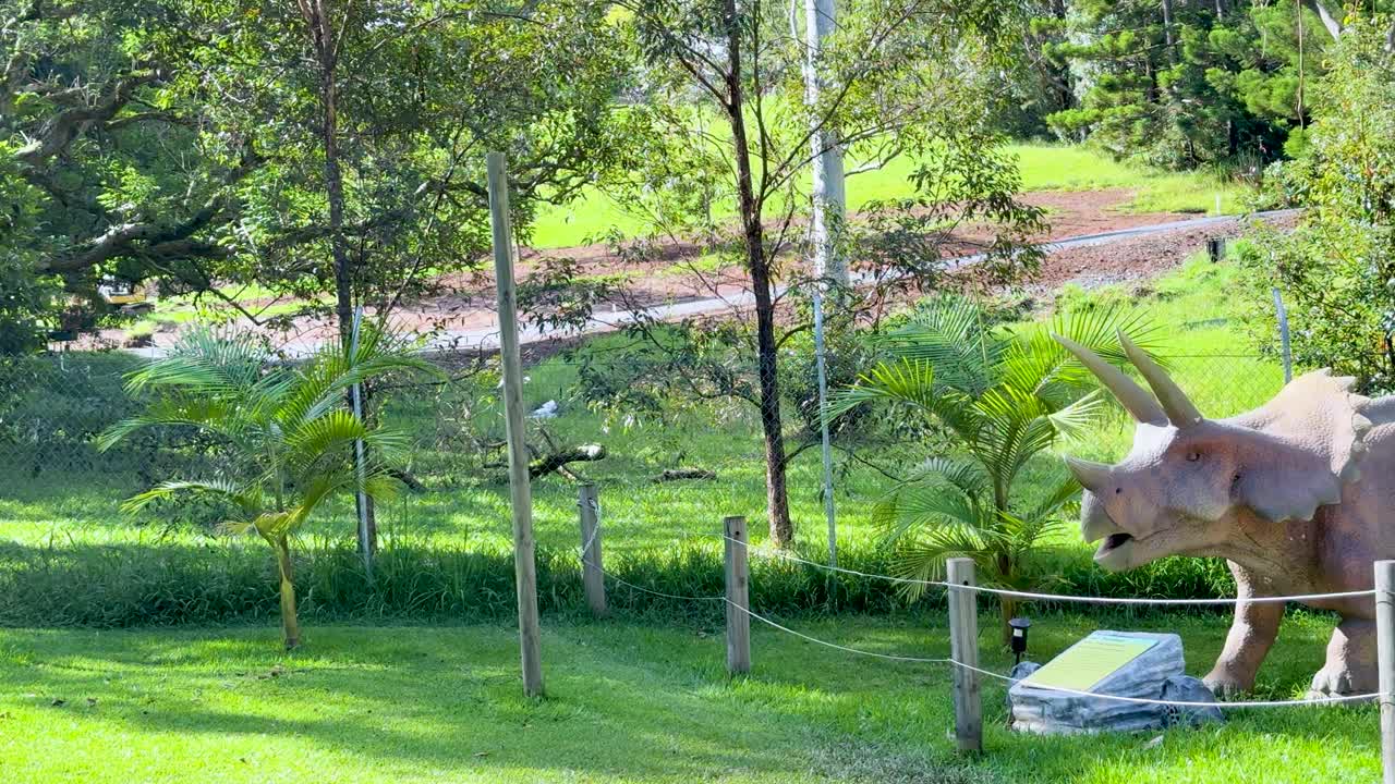 Animatronic dinosaurs move in a lush, sunlit park with steady camera panning right