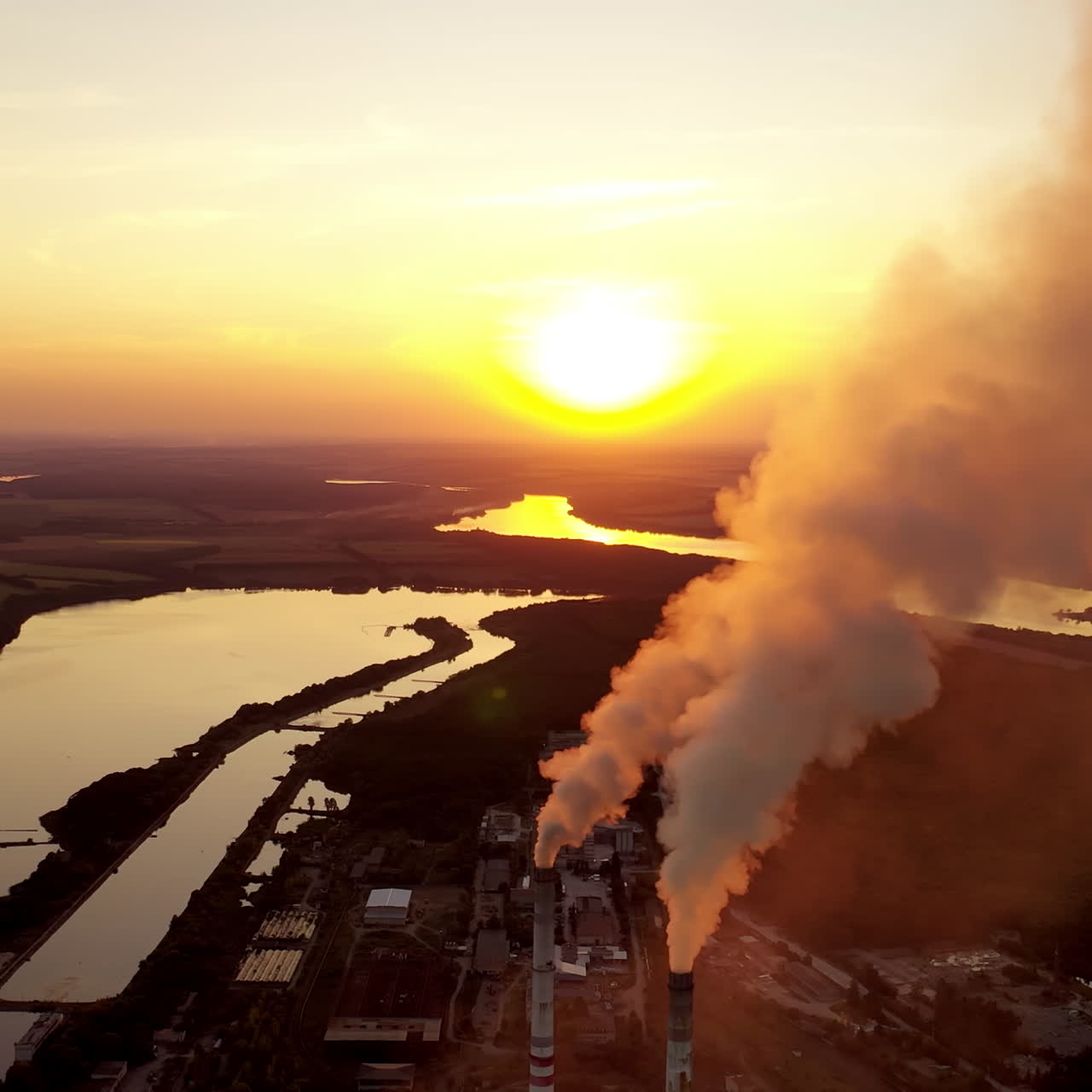 Pipes with smoke near the evening river. Pollution of the environment. Industrial plant at sunset. Dark smoke coming from plant pipes.