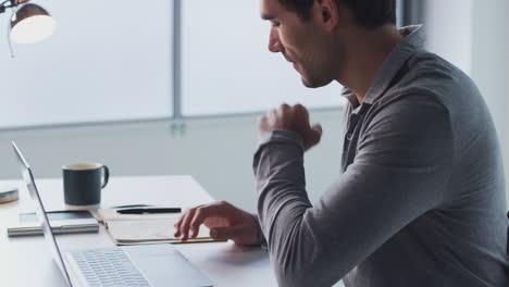 Businessman-Working-On-Laptop-At-Desk-In-Modern-Office-Looking-At-Notebook