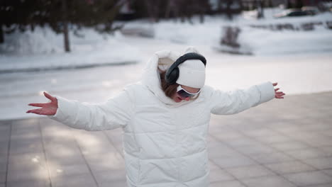 beautiful girl wearing white puffer jacket, beanie, black goggles and headphones dancing in snow covered urban park plaza, smiling and moving hands around excitedly under soft overcast winter light