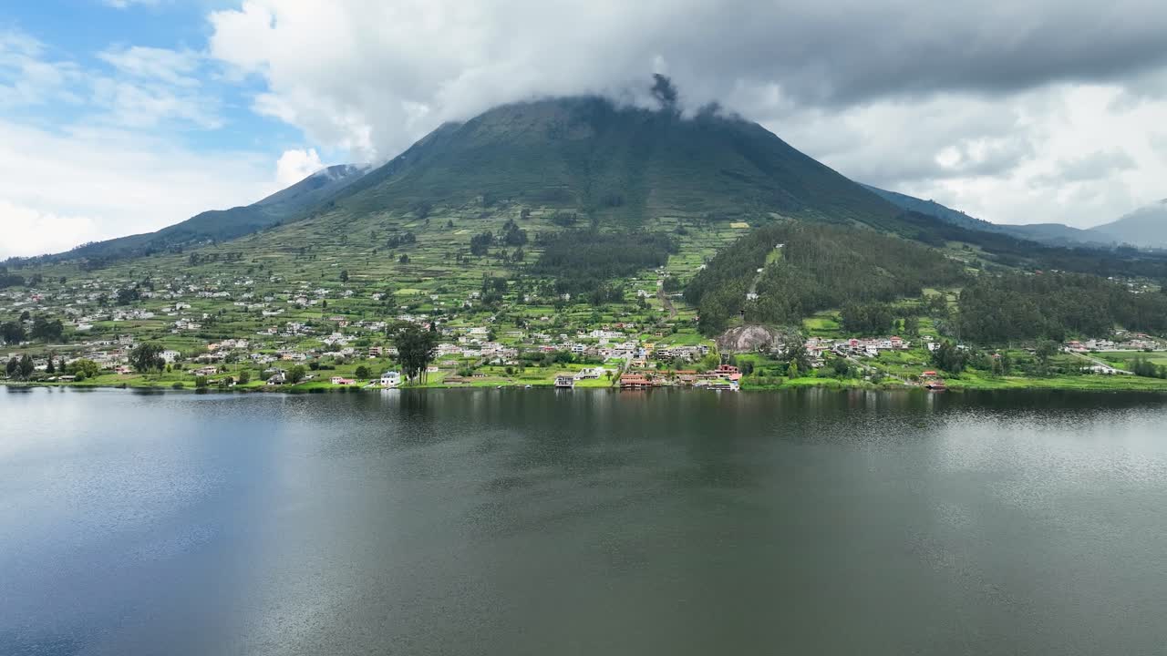 Aerial shot of a green mountain obscured by clouds at back of a town with a lagoon in front.
