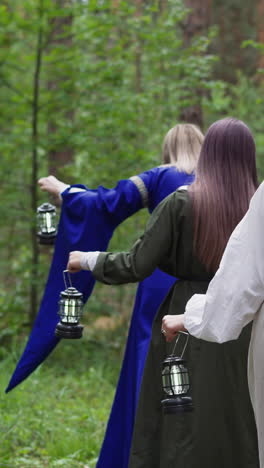 young girls in medieval long dresses walking through forest with lanterns in hands, gathering, looking for herbs