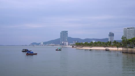 a tranquil coastal scene in ha long city showcases small fishing boats floating on calm waters, with modern high-rise buildings dotting the shoreline against a backdrop of misty mountains