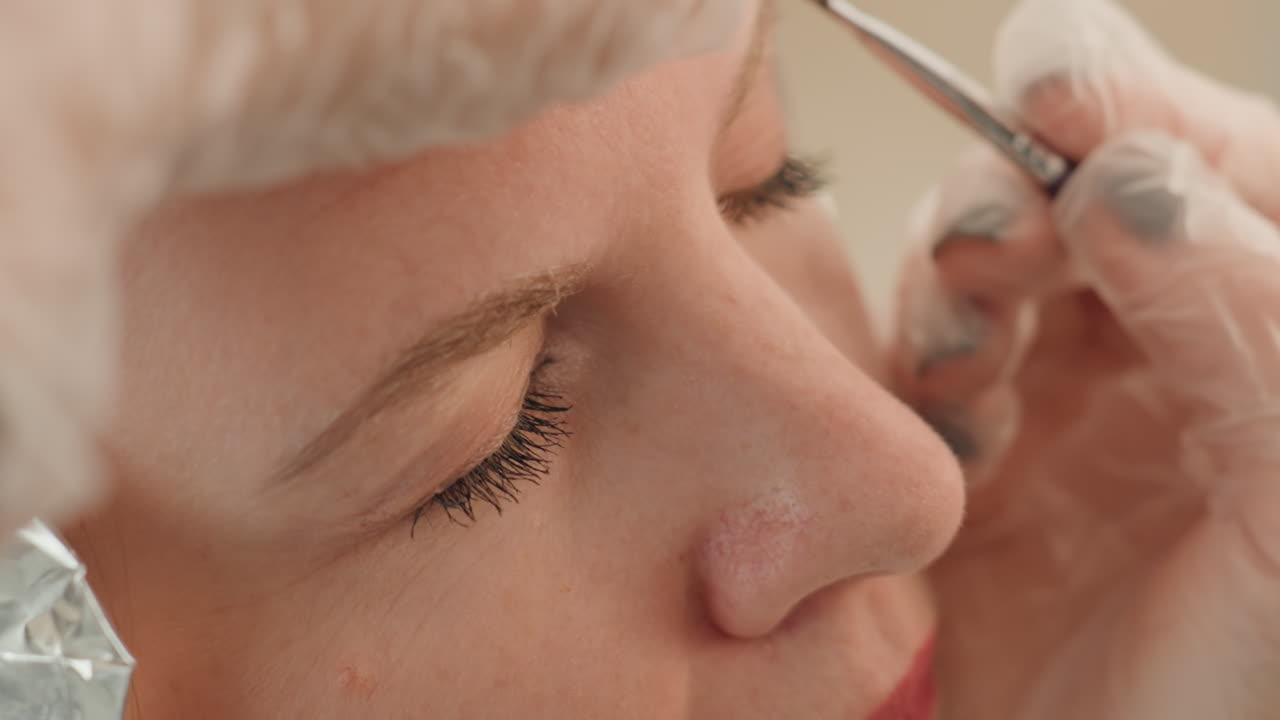 Closeup caucasian woman eyebrow microblading procedure, esthetician with gloved hands maps brow and applies pigment with precision tool, client relaxed with eyes closed, soft neutral lighting, sterile