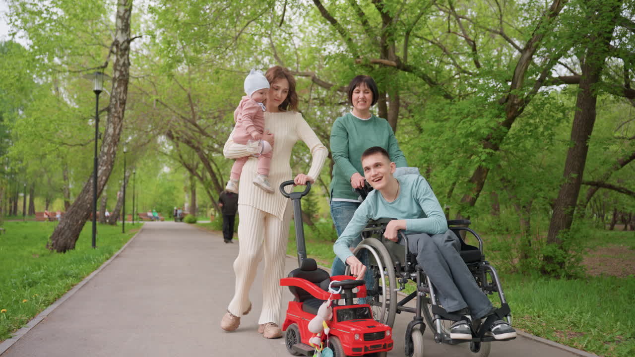 Family Walking With Wheelchair In Park Surrounded By Green Trees, Mother Holding Baby, Grandmother Smiling, Young Man In Wheelchair Inspecting Red Toy Car At Path Edge Warm Supportive Mood, Candid