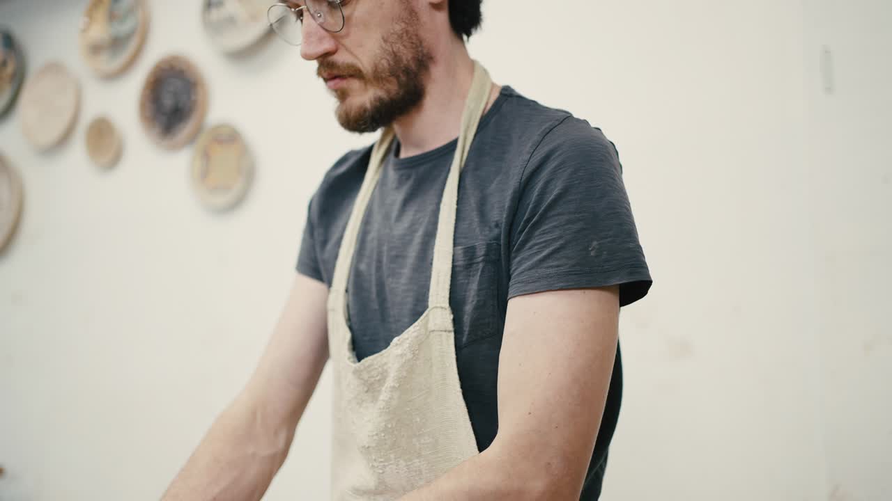 disparo de seguimiento del hombre alfarero con gafas y delantal trabajando con arcilla en la rueda de cerámica en el taller