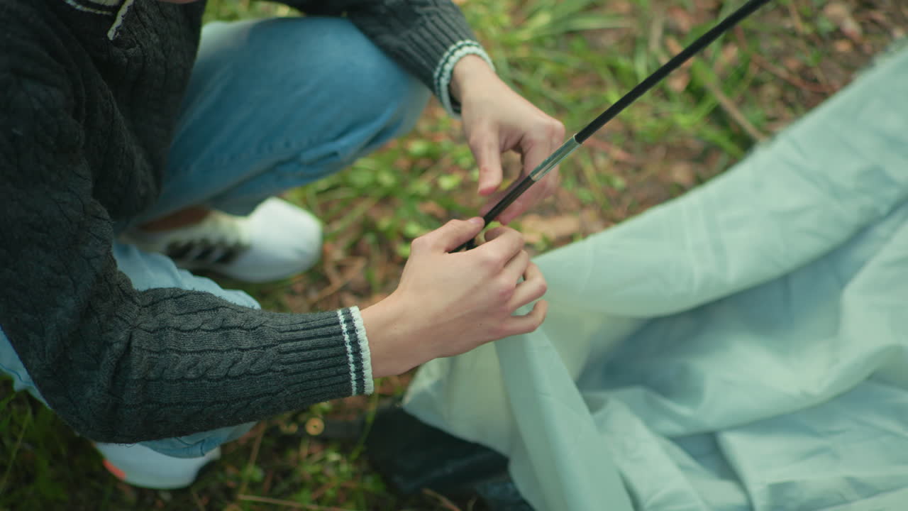 Aerial view shows person squatting in outdoor forest area adjusting flexible tent pole into fabric loop while surrounded by grass, focusing on hands and gear during camping setup