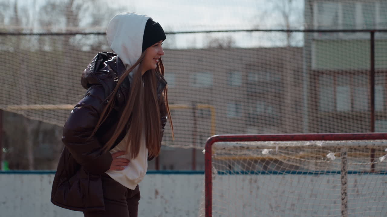 adolescente caucásico calentando cerca de la portería, con las manos en los bolsillos y caminando lentamente a lo largo de la red, chaqueta y sudadera con capucha en capas, frescor matutino y un suave telón de fondo urbano, rutina de calentamiento informal antes del entrenamiento