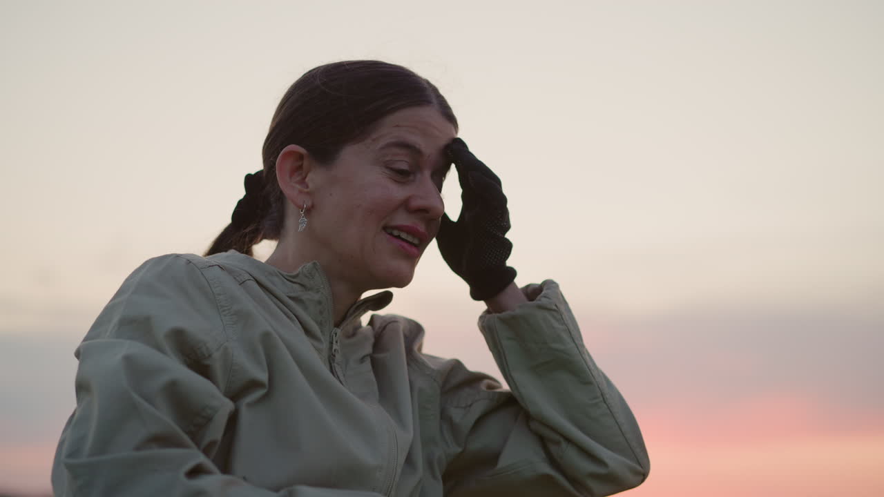 woman wiping forehead with gloved hand after hot air balloon landing in open field at sunset capturing relief and fatigue against pastel horizon in serene rural landscape