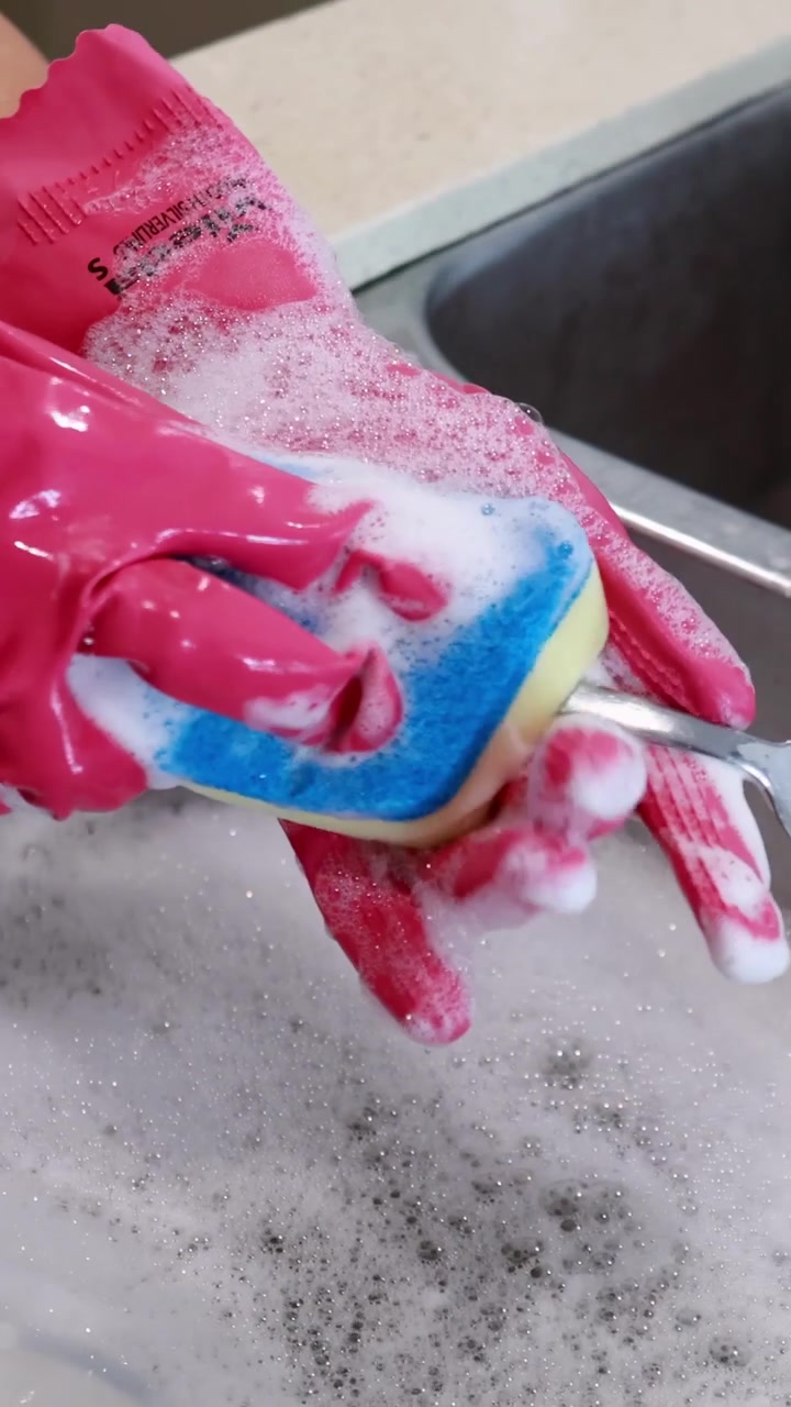 A person wearing pink gloves cleans a spoon using a soapy sponge in a kitchen sink