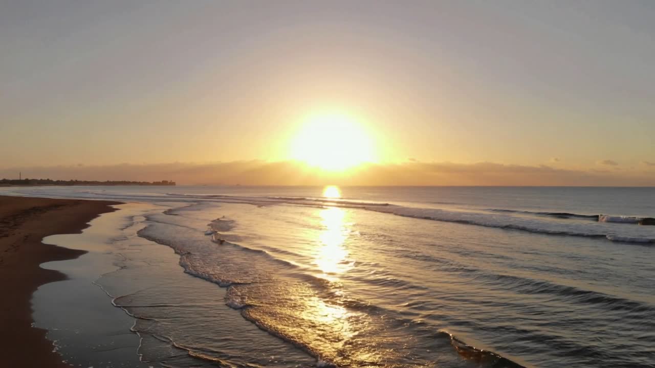 Sol nascente lançando reflexo na água ao amanhecer, revelando areia molhada curva e linhas de espuma.