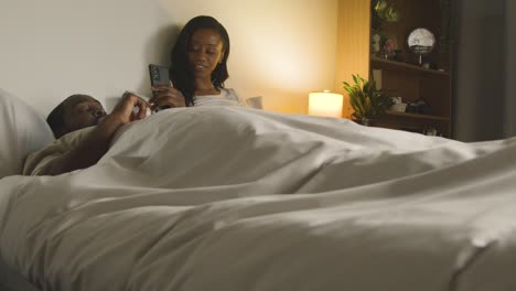 young couple at home at night lying in bed looking at mobile phones together