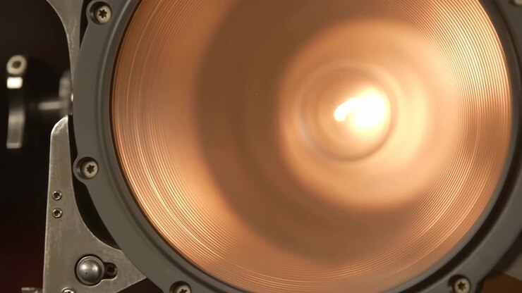 Extreme close-up of professional stage spotlight showing copper reflector, bright central filament glow, fine concentric ridges, rugged mounting hardware, warm amber light, and industrial metal detail