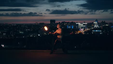 Young-blond-male-does-tricks-with-fire-breaths-fire-in-the-middle-of-the-night-with-city-skyline