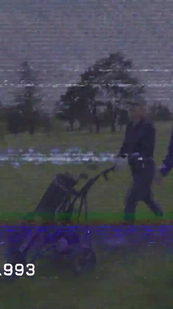 Vertikal video: 2 kvinder kommer ind fra højre, skubber en vogn på en golfbane, fortsætter runden med et 1993-overlay.