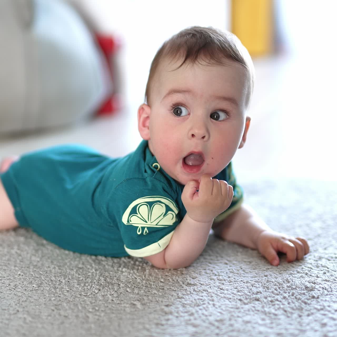 Little baby boy rolling on the floor. Cute kid opens mouth to put his fist and bangs the floor with a hand. Blurred backdrop