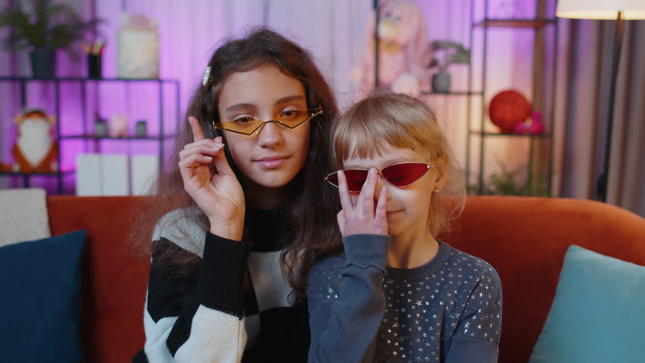 Two young girls wearing sunglasses smile and look at the camera while sitting on a couch