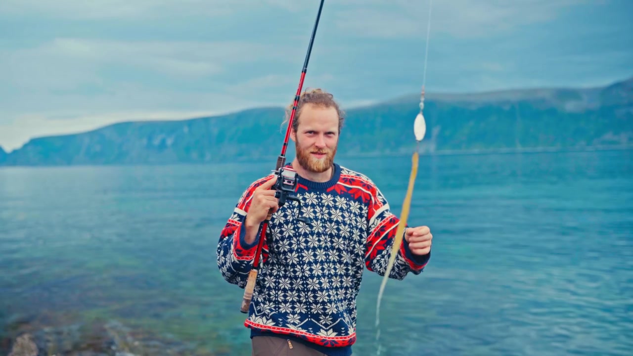 Sandland, Loppa, Norge - En mand iført en mønstret sweater holder en fiskestang ved rolige kystfarvande under en fredelig udendørs fisketur - Halvtotal